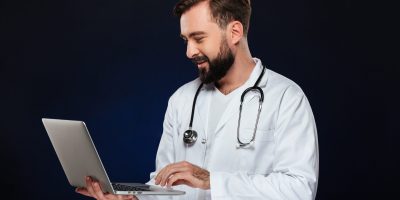 Portrait of a smiling male doctor dressed in uniform with stethoscope using laptop computer isolated over dark background