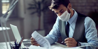 Male entrepreneur analyzing business reports while wearing face mask and working in the office during virus epidemic.